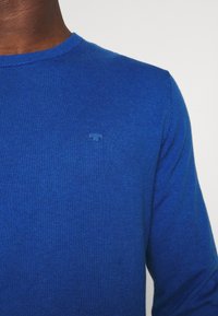 Blauer Strickpullover mit Rundhalsausschnitt. Dezenter strukturierter Stoff und kleines Logo in passendem Blau auf der Brust. Lange Ärmel.
