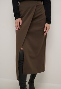 Hnědá sukně ke kolenům z jemné látky, s bočním rozparkem a zavinovacím designem. Nosená s černými koženými vysokými botami.