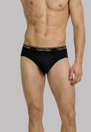 Muscular male torso wearing black Calvin Klein briefs with logo waistband, standing against a plain light gray background.