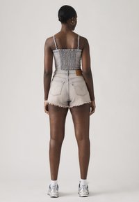 Γυναίκα που στέκεται με την πλάτη γυρισμένη, φοράει εφαρμοστή γκρι μπλούζα με κοντό μήκος και ρυθμιζόμενους ιμάντες, ανοιχτόχρωμα τζιν σορτς, λευκές κάλτσες και ασημί αθλητικά παπούτσια.