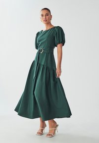 Vihreä, puffihihainen maxi-mekko, jossa on kireä vyötärö ja kultainen solki. Mekossa on kerroksellinen hameosa ja kevyt kangas.