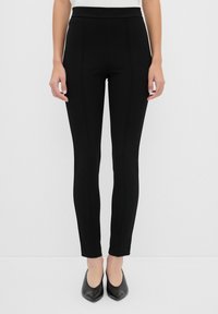 Pantalons slim noirs à texture lisse, taille haute et coutures minimales, associés à des chaussures noires à bout pointu.