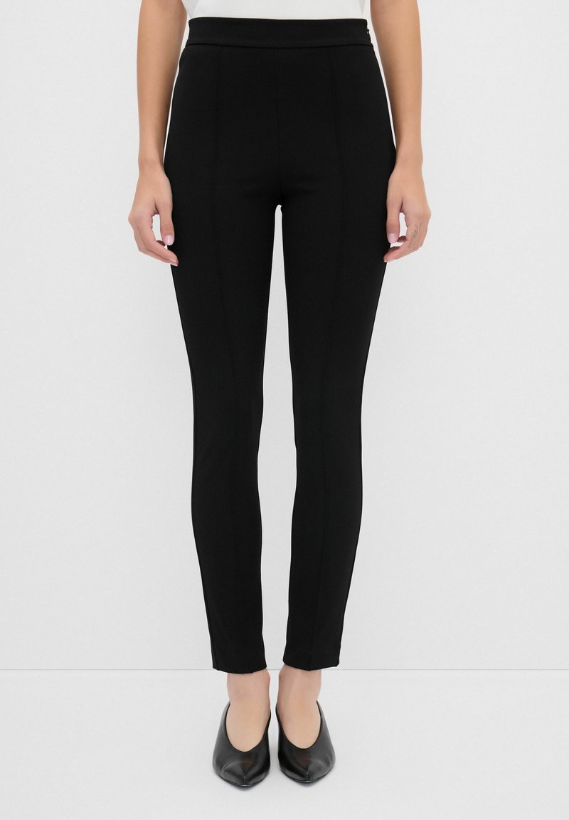 Pantalons slim noirs à texture lisse, taille haute et coutures minimales, associés à des chaussures noires à bout pointu.