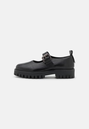 Chaussure Mary Jane en cuir noir avec semelle épaisse crantée, bride à boucle argentée et languette au talon, présentée de côté sur fond blanc.