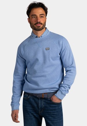 Man met donker haar en baard, gekleed in een lichtblauwe trui over een blauw patroonhemd en donkere spijkerbroek, met hand in de zak.