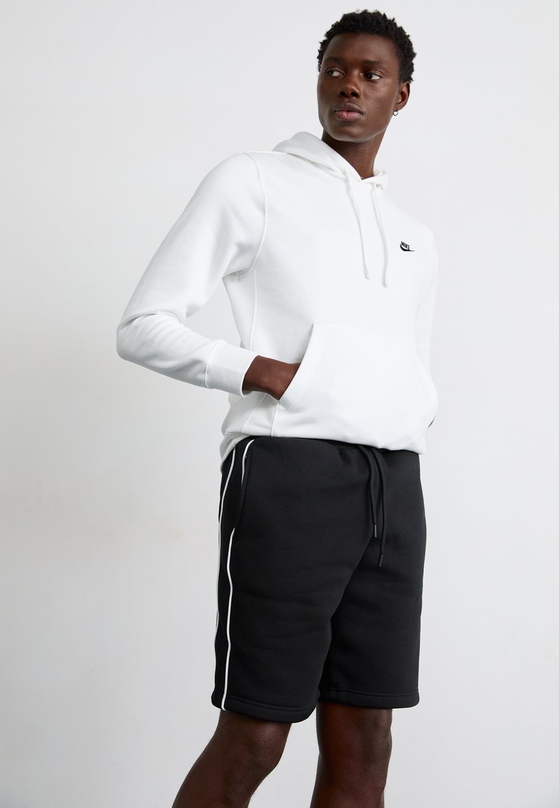 Noor mees, kellel on seljas valge Nike kapuutsiga dressipluus, käed ees taskus ning mustad lühikesed püksid valgete küljeribadega, seisab ühevärvilise tausta ees.