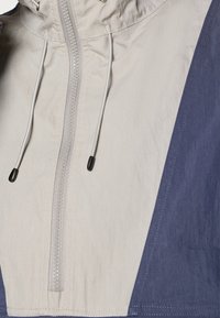 Anorak jacket in light gray and navy blue. Features a front zipper, adjustable drawstring hood, and textured, water-resistant fabric.