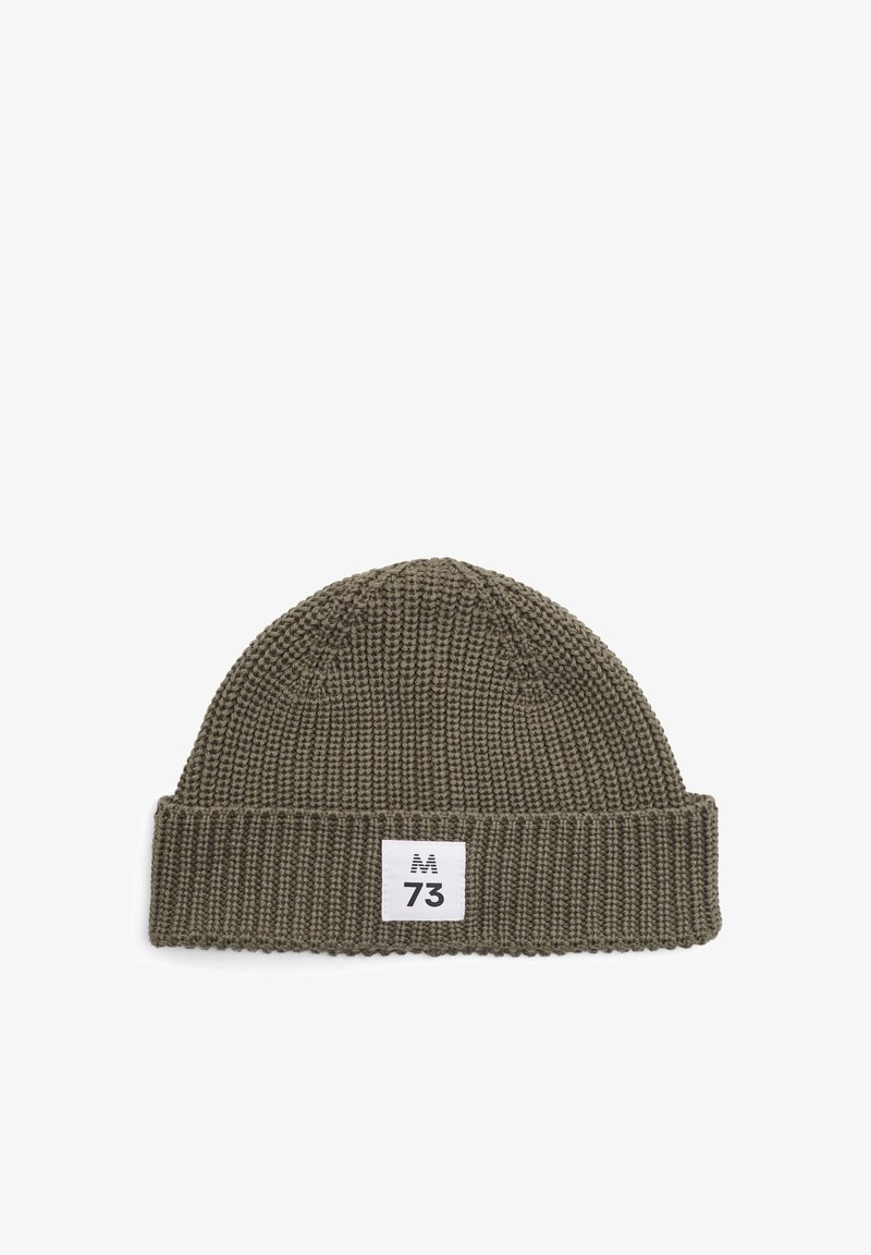 Adīta vārda cepure olīvu zaļā krāsā ar ribotu aproci un baltu etiķeti, kurā ir burti "M" un "73". Teksturēts raksts visā cepurē.