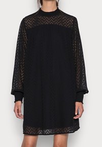 Robe noire avec manches longues transparentes, motif en zigzag texturé, col haut et coupe ample. Les matériaux en couches créent de la profondeur et un contraste dans le design.