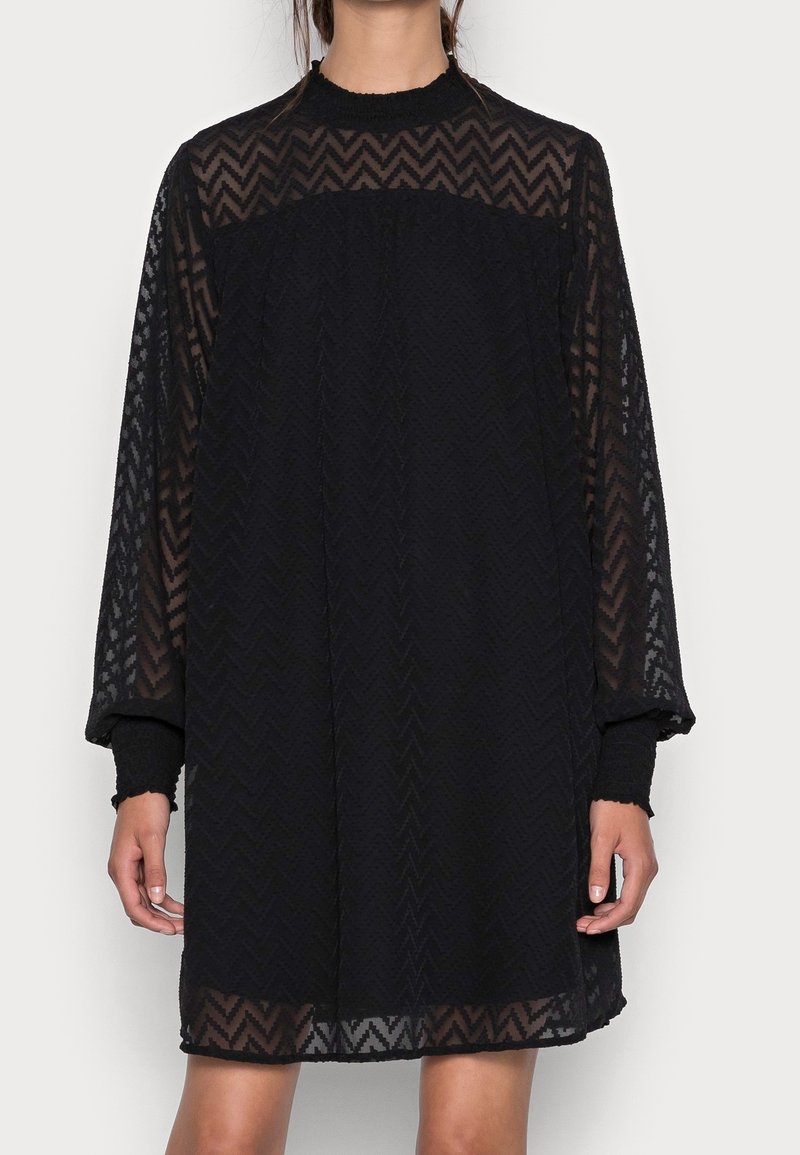 Robe noire avec manches longues transparentes, motif en zigzag texturé, col haut et coupe ample. Les matériaux en couches créent de la profondeur et un contraste dans le design.