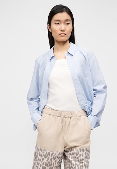 Ανοιχτό μπλε ριγέ πουκάμισο με κουμπιά και μακριά μανίκια, φορεμένο πάνω από λευκό αμάνικο τοπ, σετ με μπεζ παντελόνι με ελαστική μέση και λεοπάρ μοτίβο.