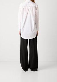 Persona in piedi con la schiena rivolta verso l'osservatore, indossa una lunga camicia bianca con bottoni e pantaloni larghi neri a gamba larga su uno sfondo bianco.