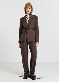 Blazer marrone su misura con vita legata, abbinato a pantaloni larghi coordinati e scarpe stringate nere. Presenta un'apertura frontale e un tessuto testurizzato.