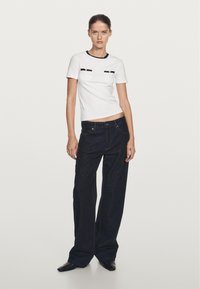 Femme debout portant un haut blanc à manches courtes avec bordure noire et un jean large foncé, posant devant un fond clair uni.