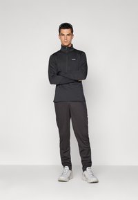 Man standing with arms crossed wearing black zip-up top, dark pants, white sneakers, and sunglasses on head against plain background.