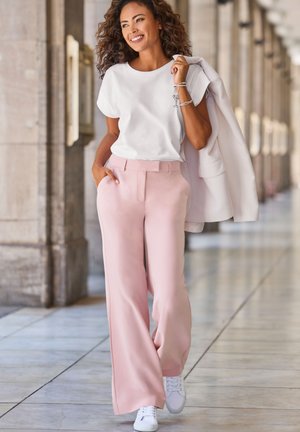Donna sorridente che cammina all'interno indossando una maglietta bianca, pantaloni rosa a gamba larga, sneaker bianche e tenendo una giacca bianca sulla spalla.