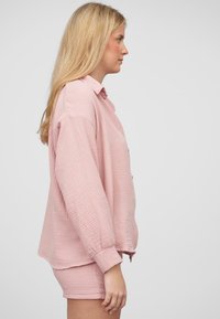 Camicia rosa pallido in misto cotone con finitura testurizzata, dotata di colletto e bottoni. Abbinata a pantaloni corti coordinati, entrambi sono morbidi.