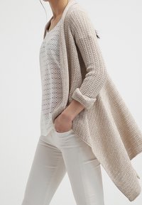 Leichte beigefarbene Strickjacke mit offenem Vorderteil, gekrempelten Ärmeln und strukturiertem Muster, getragen über einem weißen Top und hellen Hosen.