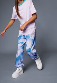 Persoon in een lichtpaarse Calvin Klein T-shirt en blauw-witte geblokte sweatpants met witte platform sneakers tegen een grijze achtergrond.