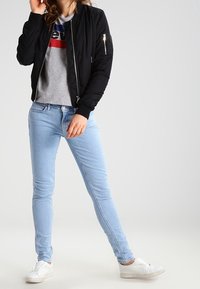 Chaqueta bomber negra hecha de un tejido ligero, con un bolsillo con cremallera, llevada sobre una camiseta gris con un logo en rojo y azul, combinada con jeans de color azul claro y zapatillas blancas.