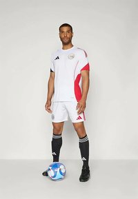 Valge jalgpallipluus punaste aktsentidega, mustad Adidas triibud ja ümmargune embleem; kokku sobitatud valgete lühikeste pükste ja mustade sokkidega. Jalgpall jalgade juures.