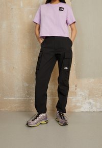 Lila kortärmad t-shirt med en svart logotyp, tillsammans med svarta cargo-byxor med sidofickor. Skorna är ljusgrå med gula sulor.