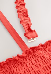 Piros elasztikus anyag, ráncolt textúrával, fodros szegélyekkel és lapos pánttal. Az arany színű csat a pánthoz van rögzítve.