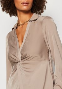 Blusa di raso beige con scollatura a V, nodo in vita e maniche lunghe. Presenta una texture morbida e liscia con dettagli sul colletto. Collana di catena dorata.