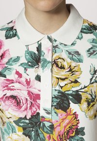 Polo floral avec un col blanc, motifs floraux vibrants en rose, jaune et teal, avec fermeture à boutons.