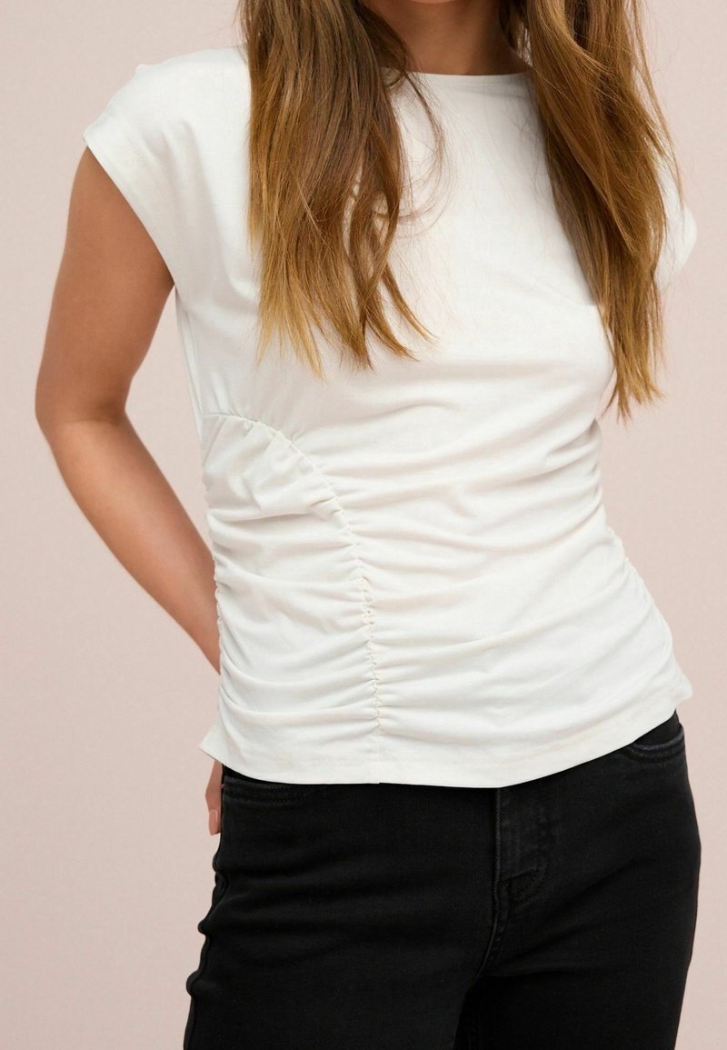 Haut blanc ajusté à manches courtes, doté de fronces latérales pour la texture et d'une finition en coton lisse. Porté avec un jean noir.