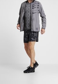 Szara kurtka bomber z nadrukami graficznymi, szary t-shirt w wzory, czarne spodenki z różnorodnymi wzorami oraz czarne buty sportowe.