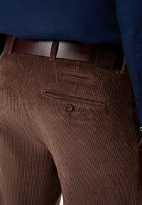 Gros plan sur un pantalon en velours côtelé marron avec une poche arrière boutonnée et une ceinture en cuir foncé, porté avec un top bleu marine.