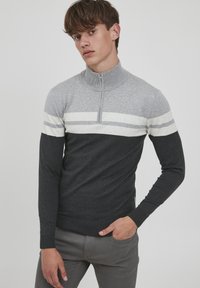 Grå zip-genser med en kontrastfarge i mørkegrå nederst, med hvite horisontale striper. Laget av et glatt, teksturert stoff.