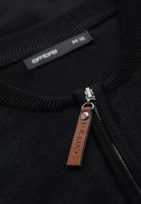 Sweat à zip noir avec un col côtelé et une tirette en cuir gravée du nom de la marque. Tissu texturé visible.