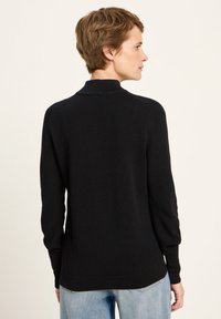 Frau mit kurzen braunen Haaren, die einen schwarzen Langarmpullover und hellblaue Jeans trägt, steht vor einem einfarbigen hellen Hintergrund.