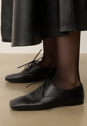 Schwarze Leder-Oxford-Schuhe mit spitzem Zeh, minimalistischem Schnürung und elegantem Design, kombiniert mit schwarzen, transparenten Strumpfhosen.