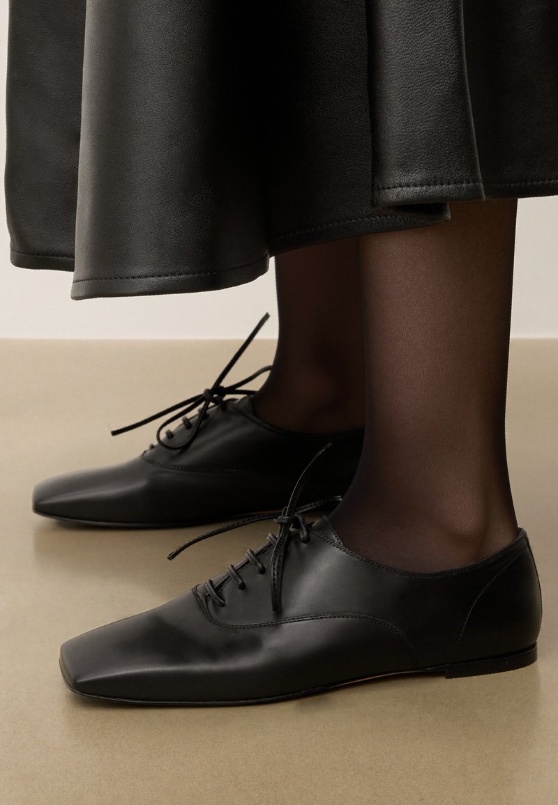 Svarta läderskor av oxfordmodell med spetsig tå, med minimalistiska snörningar och en smidig design, tillsammans med svarta genomskinliga strumpor.