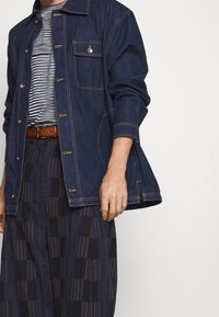 Una persona que lleva una chaqueta de mezclilla oscura sobre una camisa a rayas, con pantalones marrones de patrón a cuadros, de pie contra un fondo liso.