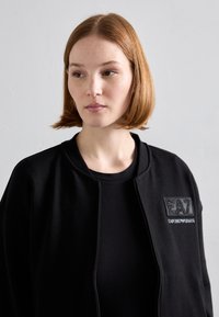 Crna jakna sa zatvaračem na prednjem dijelu, okruglog vrata, izrađena od glatkog materijala, ima teksturiranu zakrpu s EA7 logom na lijevoj strani prsiju.