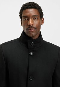 Cappotto in misto lana nera con colletto alto, chiusura a bottoni e texture liscia. Presenta due bottoni neri nella parte anteriore.