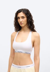 Γυναίκα με σγουρά σκούρα μαλλιά, φορώντας λευκό αθλητικό σουτιέν Calvin Klein και ανοιχτόχρωμο παντελόνι, σε απλό λευκό φόντο.