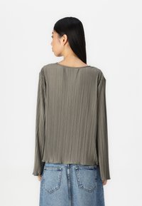 Langärmlige, plissierte Bluse aus graugrünem Stoff. Verfügt über einen runden Ausschnitt und eine lockere Passform, kombiniert mit einem Jeansrock.