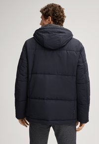 Giubbotto imbottito blu scuro con cappuccio, caratterizzato da un design trapuntato, cuciture verticali e polsini elasticizzati. Texture liscia, stile minimalista.