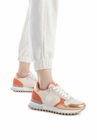 Zapatillas en blanco y melocotón con patrones texturizados, detalles metálicos en oro rosa y cordones blancos. Suela de goma con elementos de tracción.