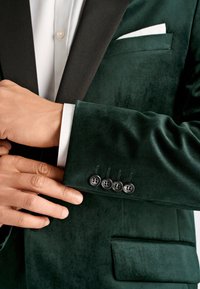 Hands adjusting the cuff of a dark green velvet blazer with four black buttons, white shirt cuff visible, and white pocket square.