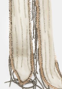 Accessoire en perles présentant des bandes de tissu allongées en beige, agrémentées de perles métalliques et transparentes, créant un effet texturé et stratifié.