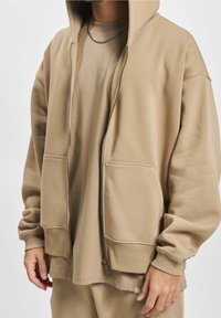 Beige Kapuzenpullover mit Reißverschluss, Kapuze, Kängurutaschen und lockerer Passform. Weiche Textur und ein passendes beiges T-Shirt darunter.