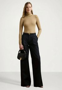 Mulher vestindo um pulôver bege de gola alta, calças largas pretas de cintura alta, segurando uma pequena bolsa preta, em pé contra um fundo simples.