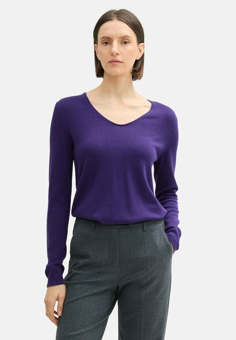 Lila langärmliger V-Ausschnitt-Pullover aus weichem Material, kombiniert mit grauen gemusterten Hosen. Der Pullover hat ein tailliertes Design und eine lockere Silhouette.