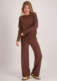 Bruin gebreid topje met brede halslijn, gecombineerd met bijpassende wijde broekspijpen. Beide hebben een zachte textuur en een taille met trekkoord. Licht schoeisel met strepen.